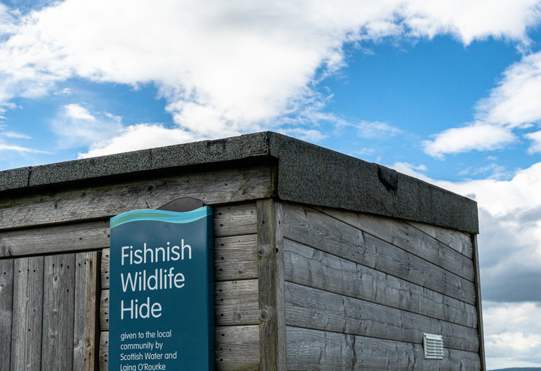 The top of a wooden wildlife hide with a blue sign saying 'Fishnish Wildlife Hide' and blue sky behind