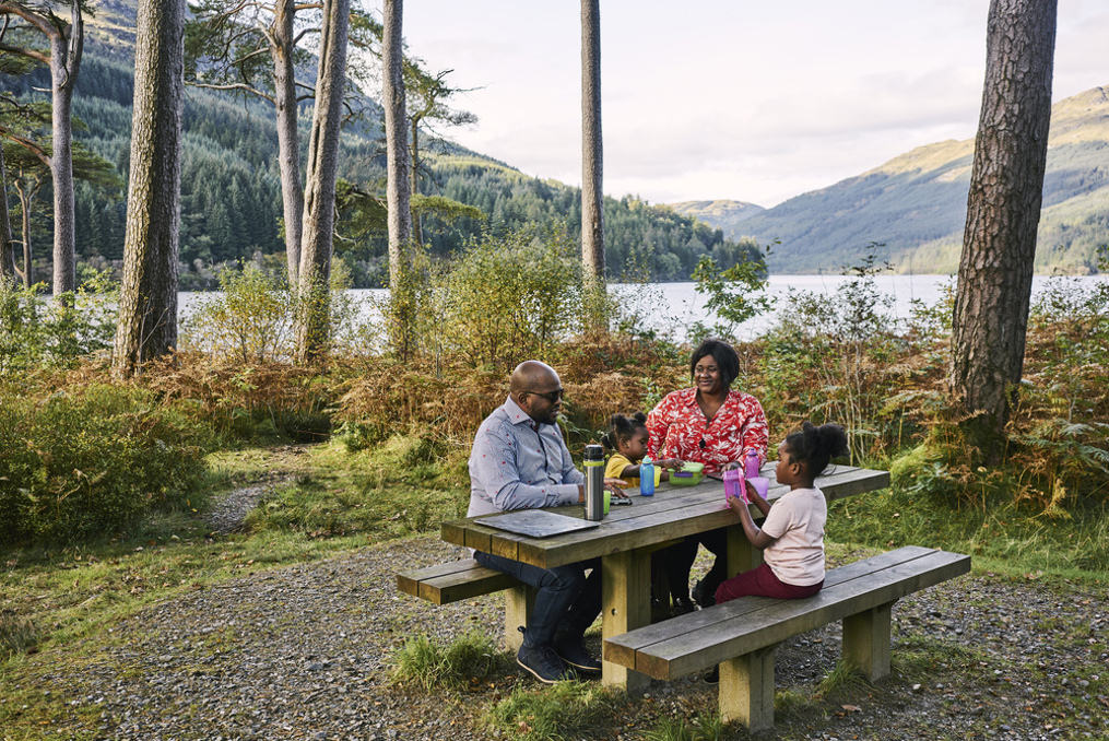 Man, woman and two young girls sit at picnic table with food and drink, at Jubilee Point, on shore of Loch Eck