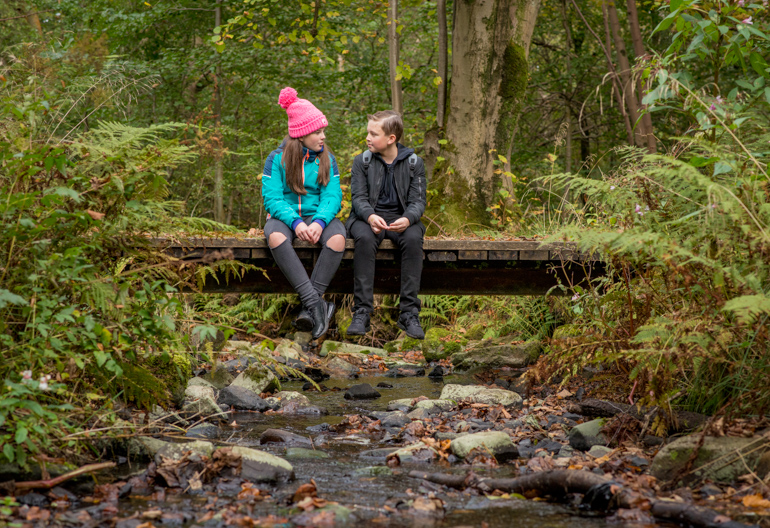 Young girl and young boy sit together on wooden bridge over stream, Bluebell Wood, Renfrewshire Woods, near Johnstone