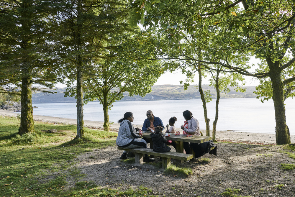 Family sit at picnic table under trees with loch in the background.