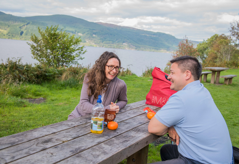 Woman and man sit at picnic bench with a snack, at Change House, south side shore of Loch Ness