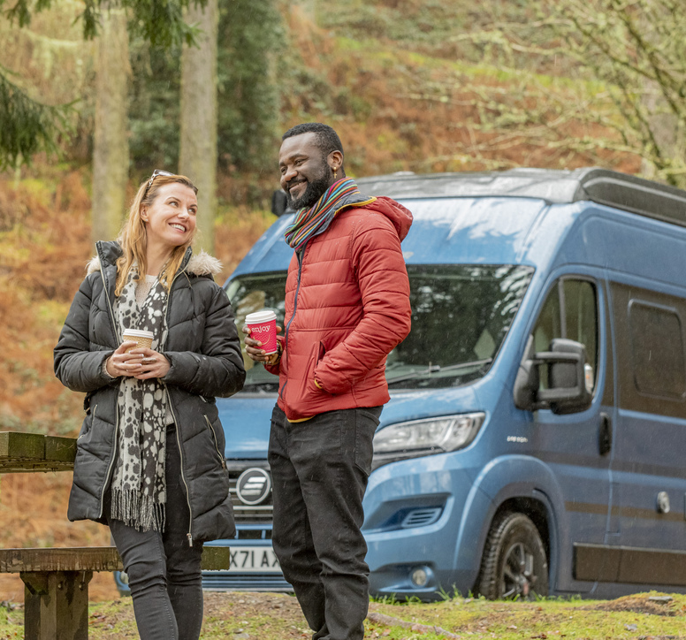 Two people enjoying a coffee next to a picnic table with their camper van behind them