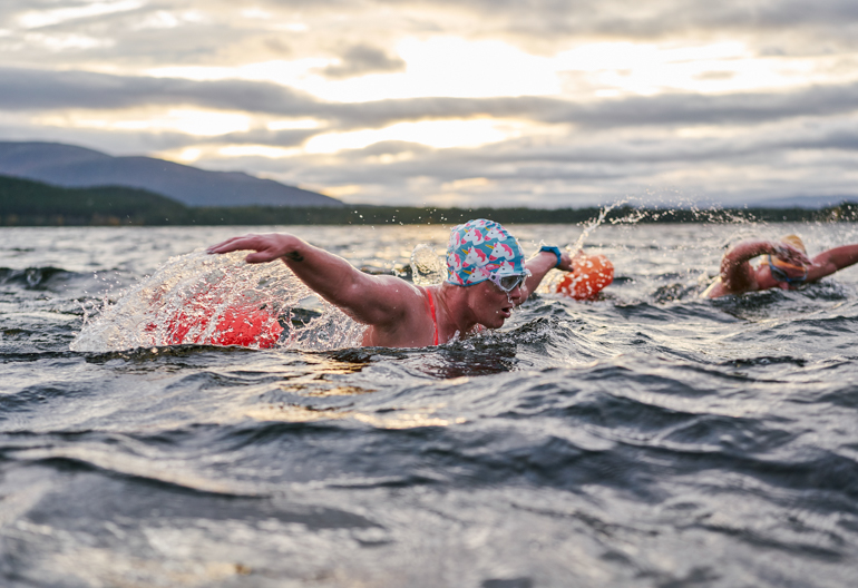 Woman in colourful skullcap swimming mid-stroke in loch under a cloudy sky.