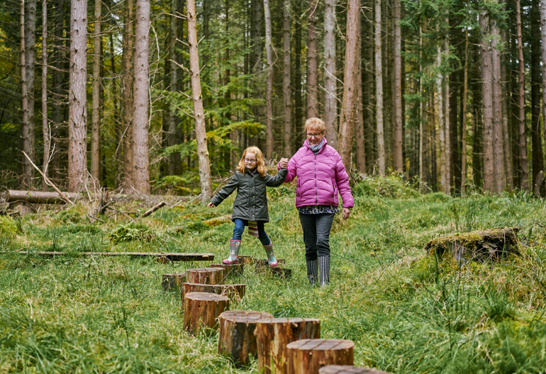 Woman holding hand of child who is stepping from one tree stump to another in middle of forest.