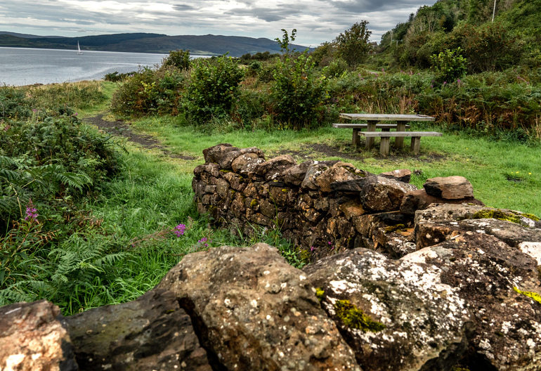 Start of a path way with signage and a picnic table and water in the background