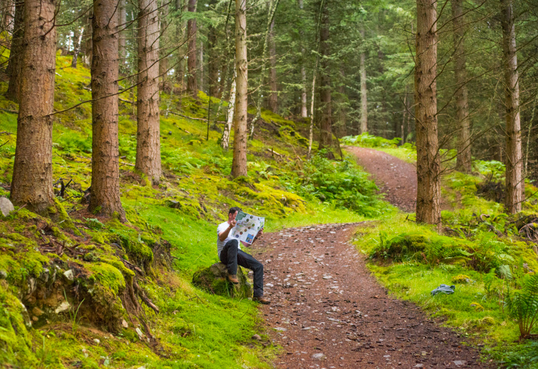 Man sits on boulder on woodland trail and consults informational leaflet, Craigmonie Forest, near Drumnadrochit