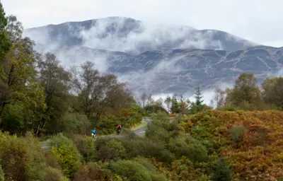 Two cyclists riding gently uphill on a road bordered by green trees and looking out to a large bare hillside with wisps of cloud.
