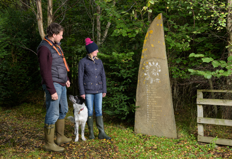 Man and woman, with dog, look at the Gaelic stone sculpture at entrance to the Tree Alphabet, Borgie Breco, Borgie, Sutherland