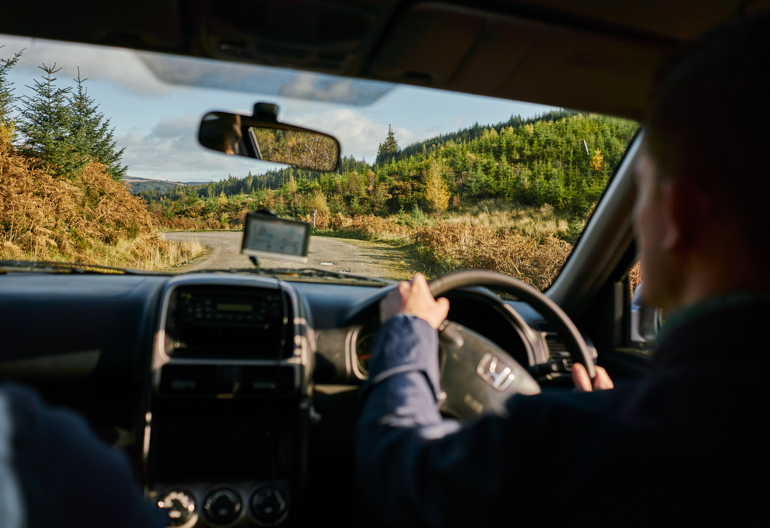 A man inside a car, driving down a forested road with a young woodland in the distance