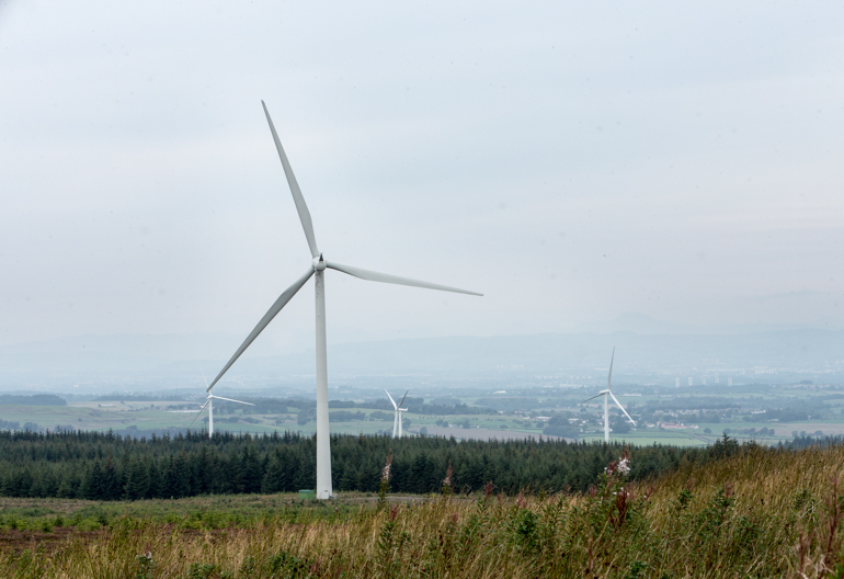Large windmills in a windfarm with trees and fields.